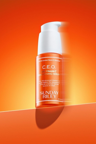 A high-impact product hero shot of a brightening serum bottle centered against a vibrant orange background, utilizing clean geometry and bold color blocking.